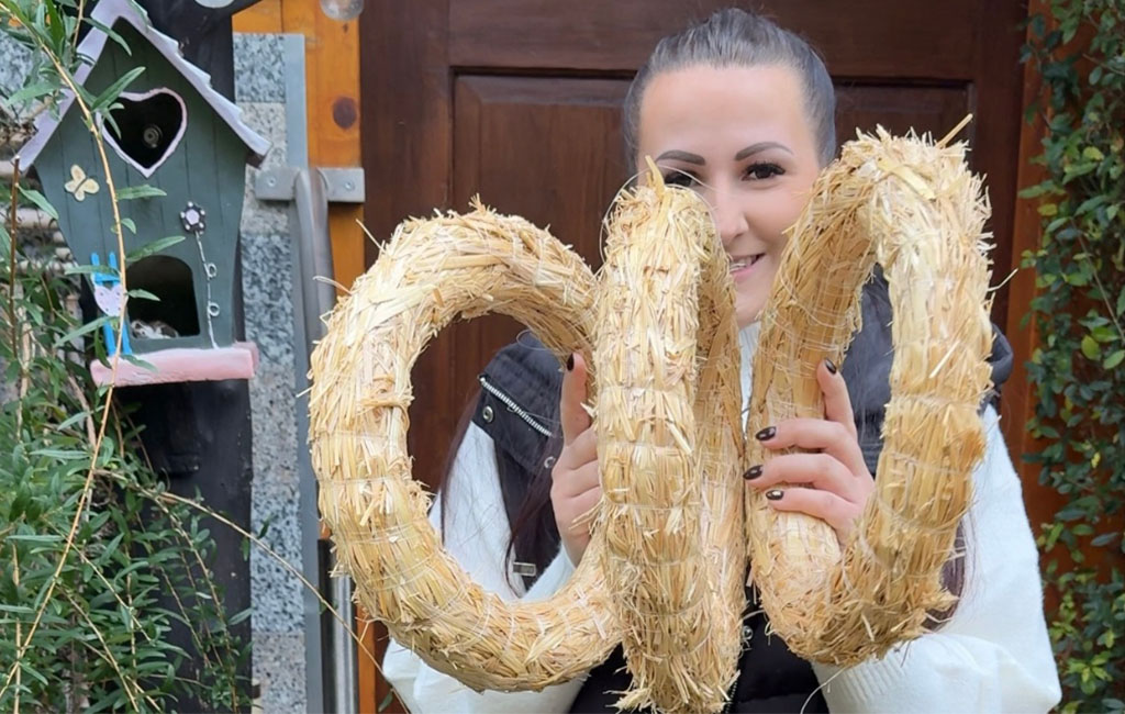 A woman holds three large straw wreaths in her hands, smiling from behind the crafting material.