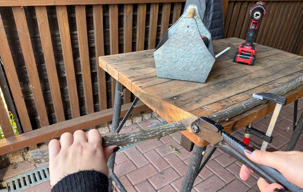 A person cuts a branch with a handsaw on a table; the branch is clamped in place.