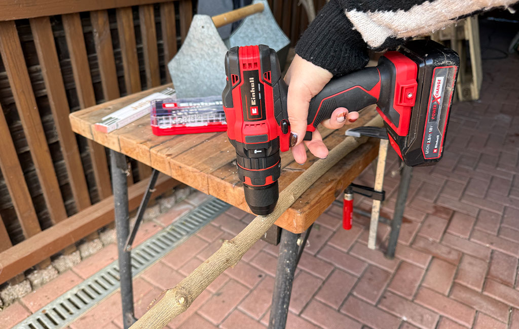 A person drills a hole into a branch using an Einhell cordless drill driver.