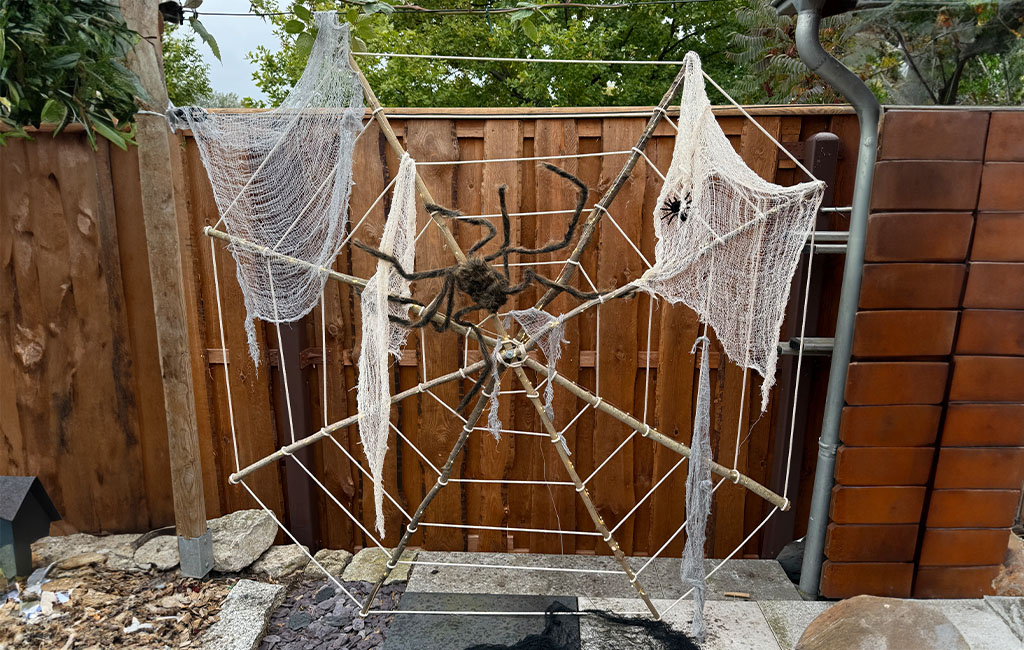 Large spider web made of ropes with white cloth and two artificial spiders, mounted on a wooden frame in a garden.