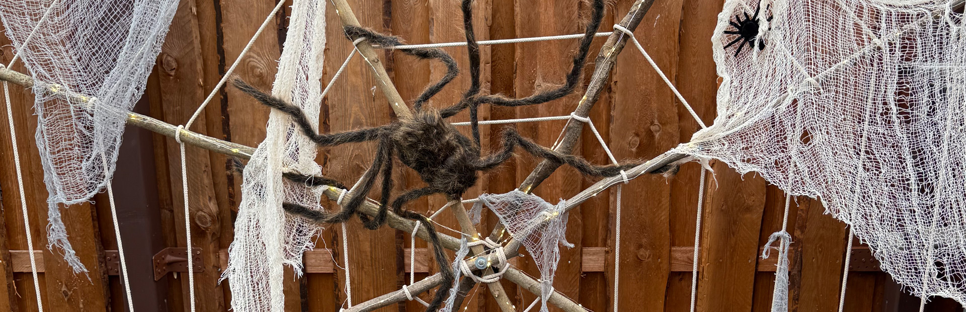 Large spider web made of ropes with white cloth and two artificial spiders, mounted on a wooden frame in a garden.