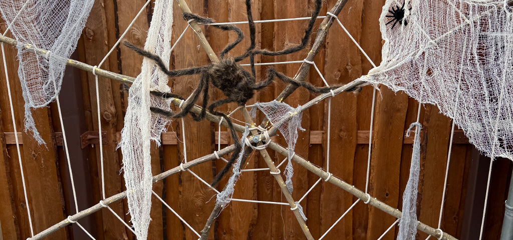 Large spider web made of ropes with white cloth and two artificial spiders, mounted on a wooden frame in a garden.