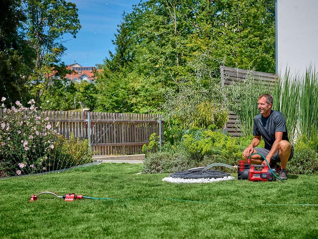A man kneeling next to cordless garden pump which pumps water to a lawn sprinkler. 