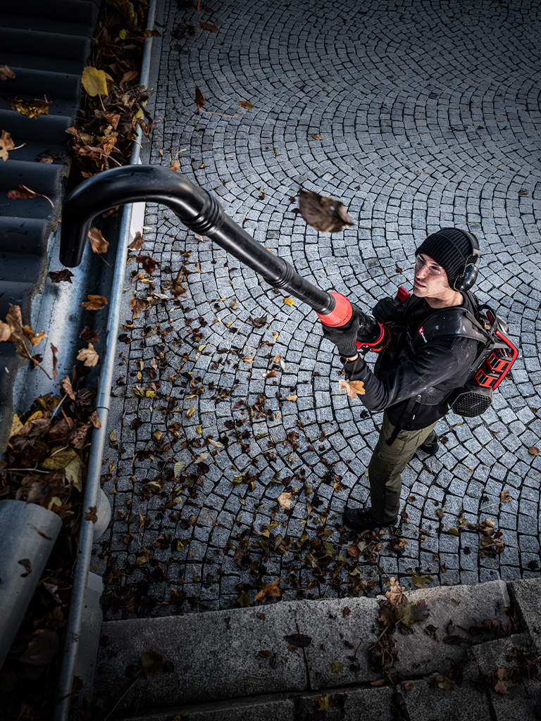 A man uses an Einhell cordless leaf blower with a long gutter cleaning attachment to clear leaves from a gutter.