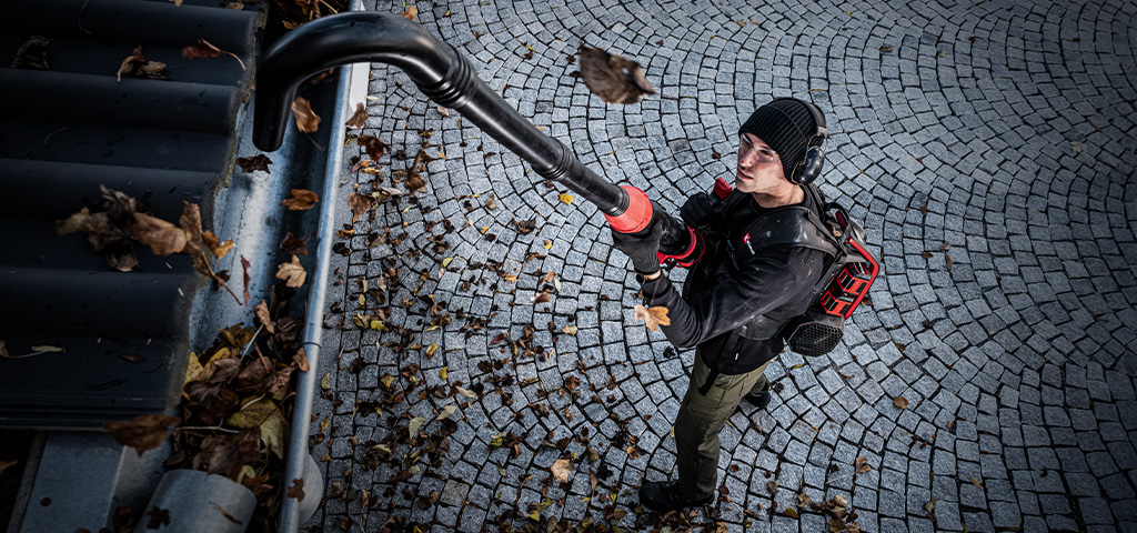 A man uses an Einhell cordless leaf blower with a long gutter cleaning attachment to clear leaves from a gutter.