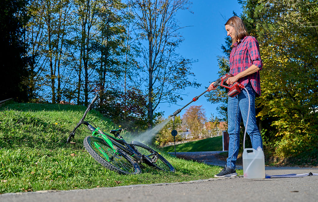 A woman in a checkered shirt and jeans is cleaning a bicycle with a cordless pressure washer outdoors in sunny weather.