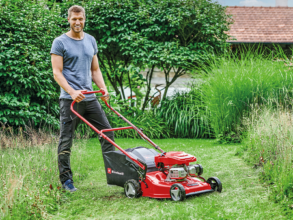 Ein Mann steht mit einem Benzin-Rasenmäher von Einhell auf abgemähtem Rasen.
