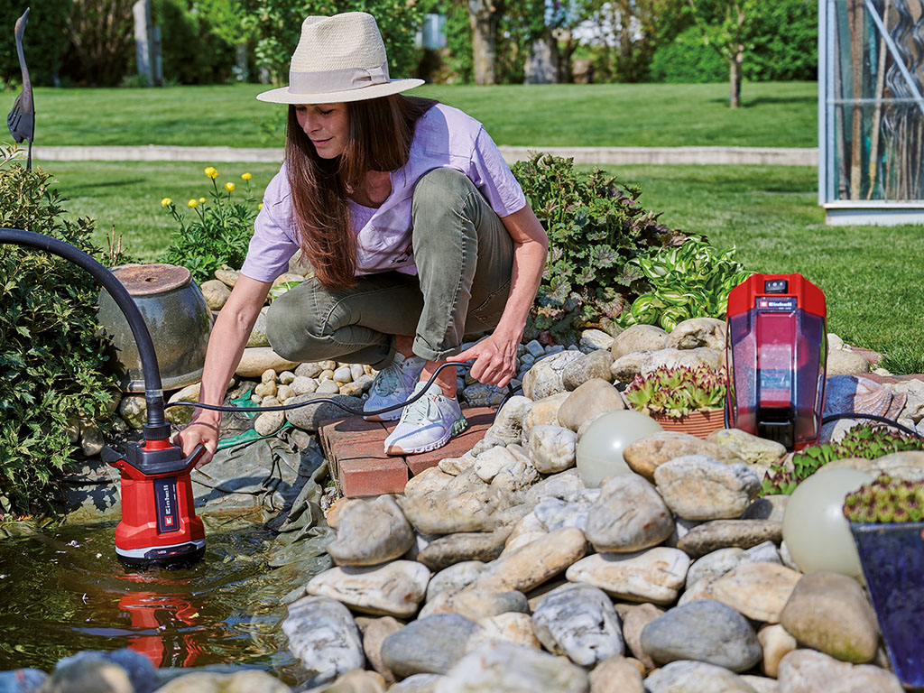 A woman uses an Einhell dirty water pump to vacuum water from a garden pond beside a pile of stones.