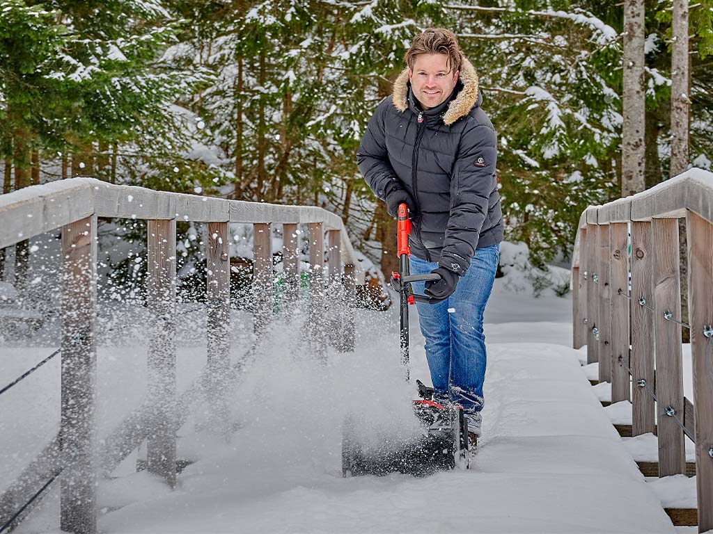 Man clearing snow with a cordless snow blower on a snow-covered bridge in front of a forest
