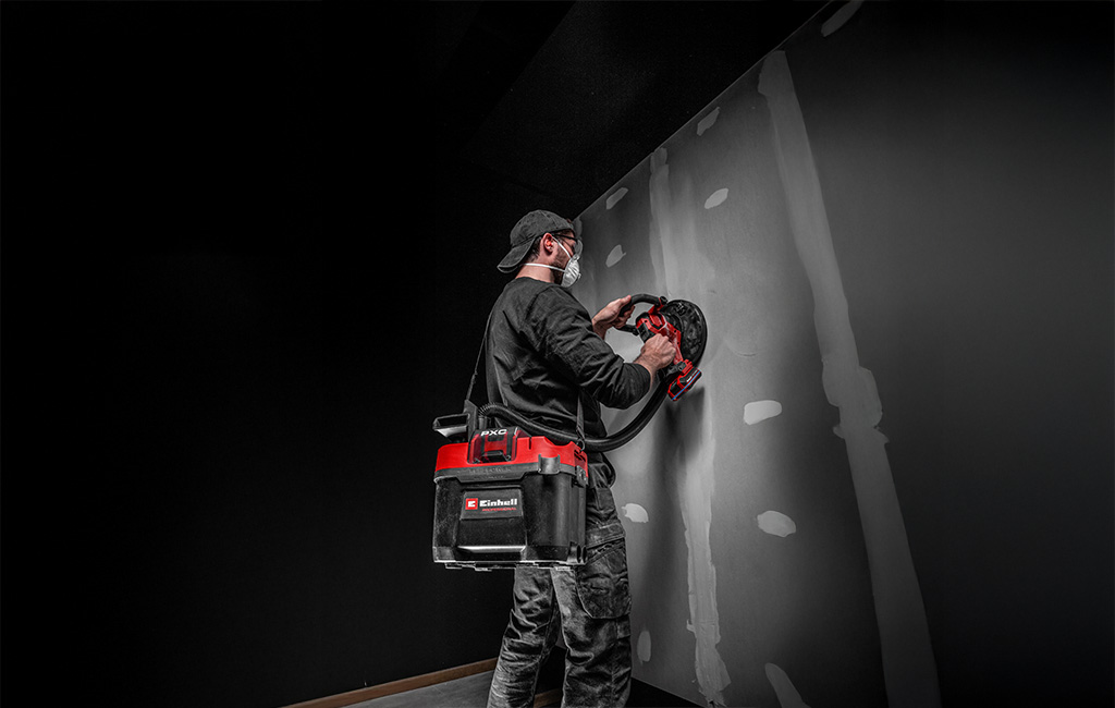 A person vacuums along a wall with a cordless vacuum cleaner, with visible dust particles being sucked in.