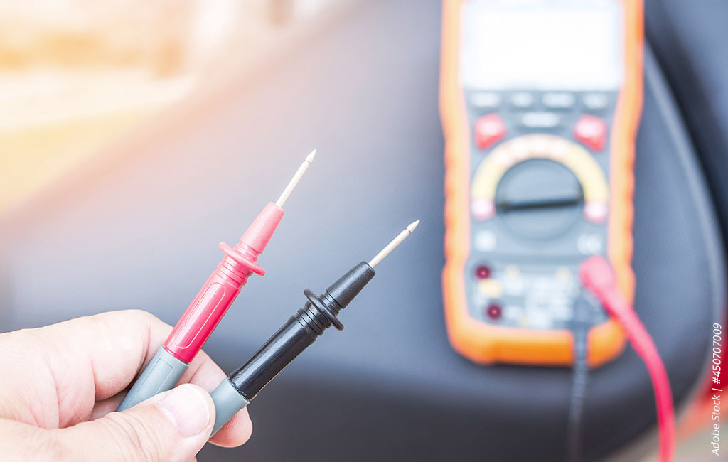 Hand holding two test probes in front of a multimeter to check voltage or battery.