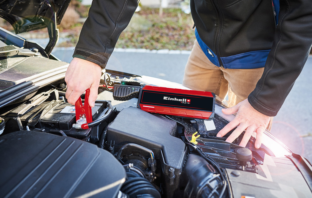 Einhell battery jump starter connected to a car battery in the engine compartment.
