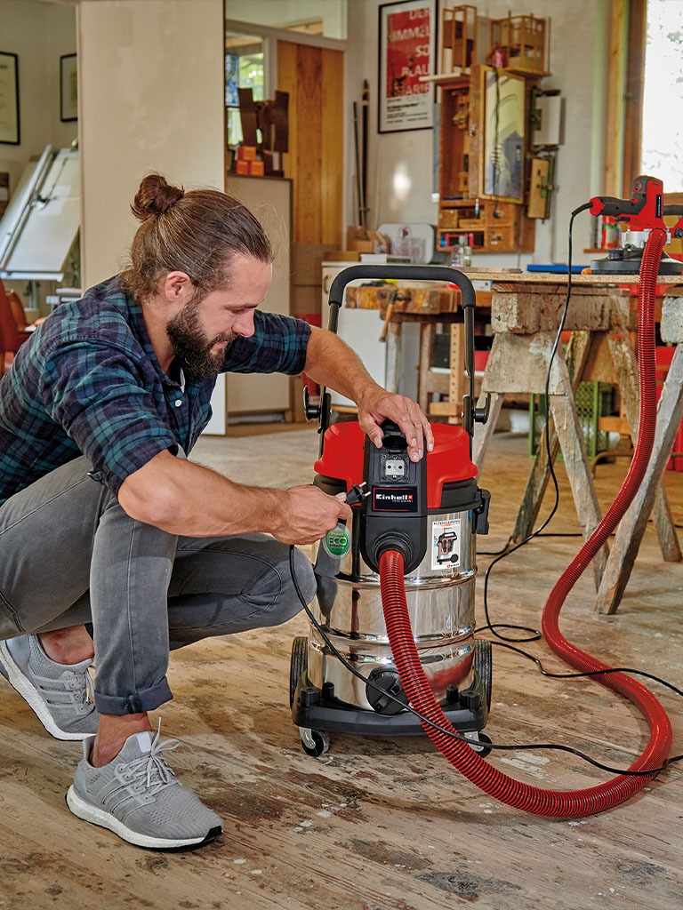 A man uses an Einhell wet and dry vacuum cleaner while working at a workbench.
