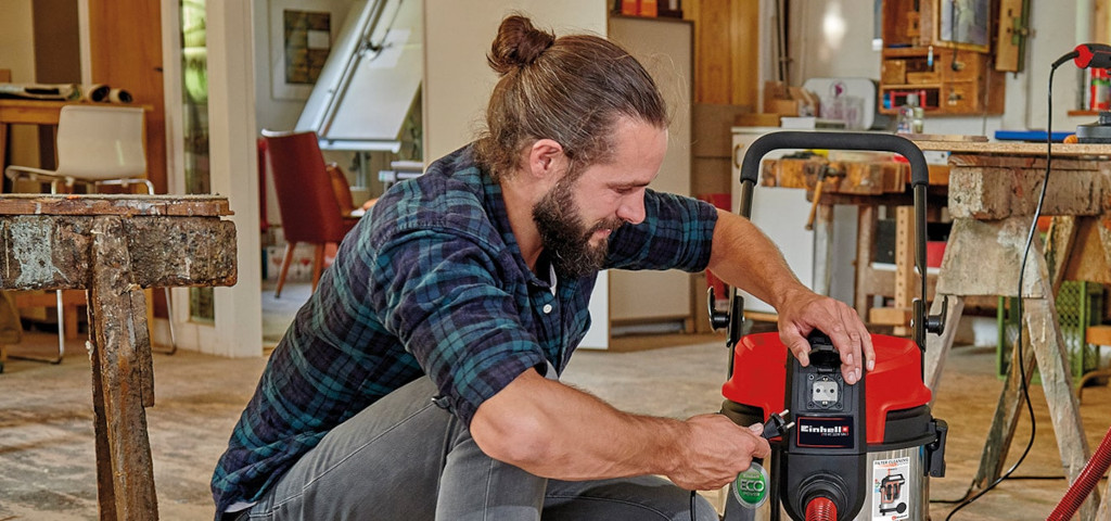 A man uses an Einhell wet and dry vacuum cleaner while working at a workbench.