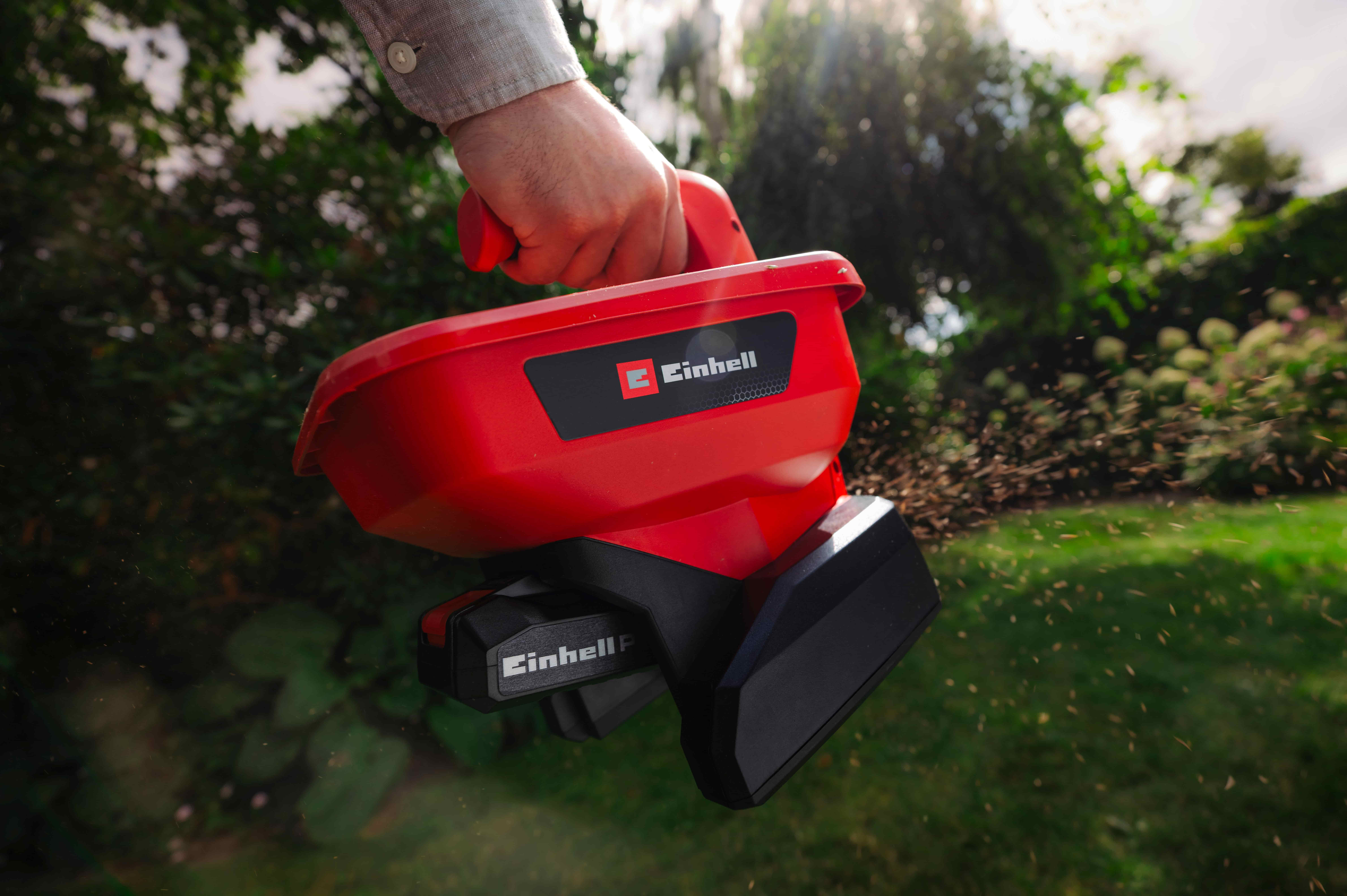 Hand streut Saatgut aus einem roten Einhell Akku-Streuer, während kleine Samen über den Garten fliegen.