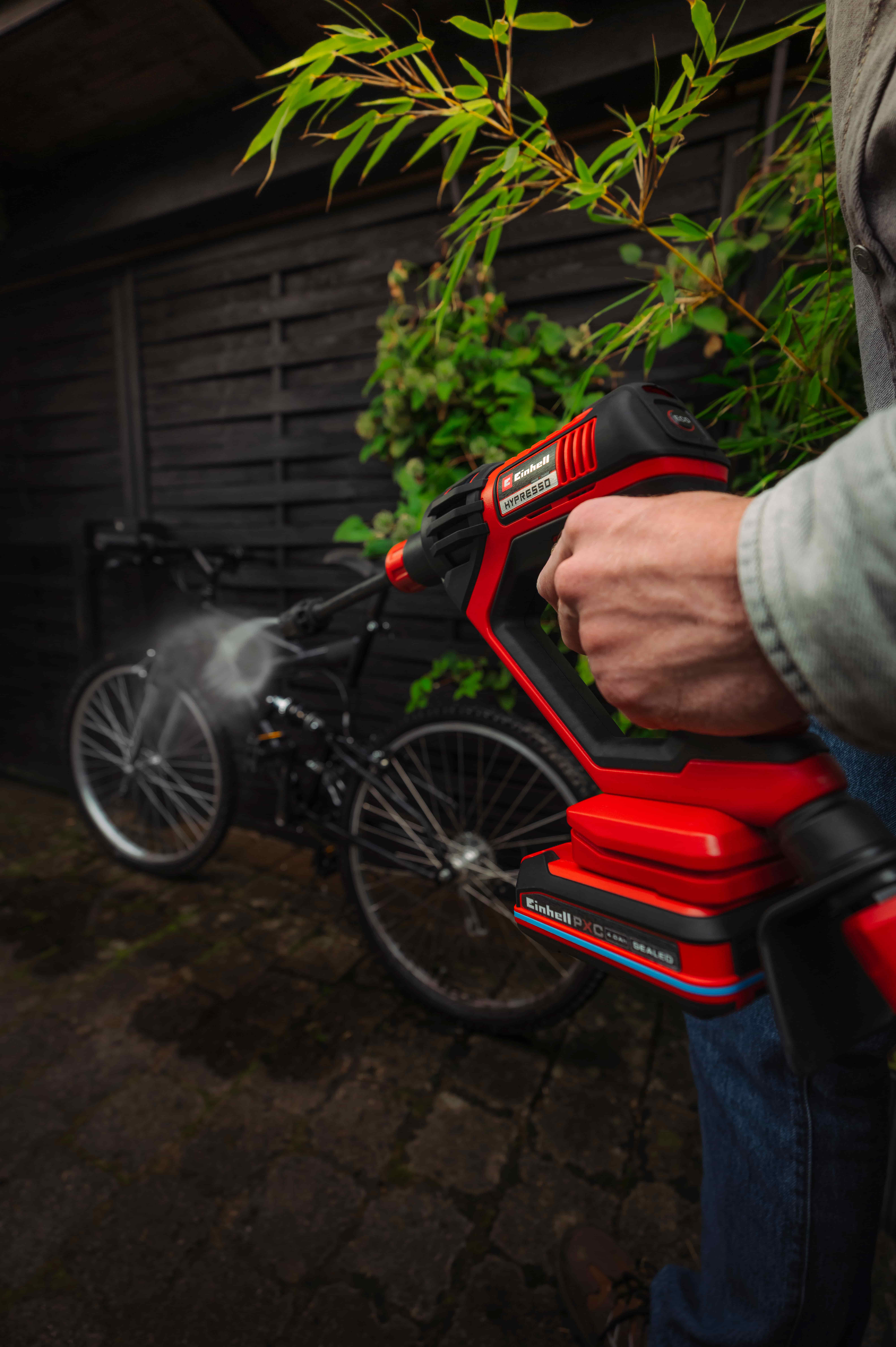 Person reinigt ein Fahrrad mit einem roten Einhell Akku-Hochdruckreiniger im Hof neben einer Holzwand.