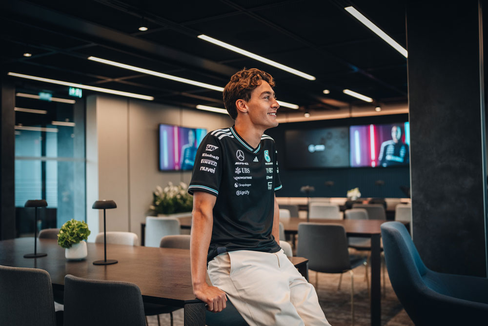 George Russell, pilota del team Mercedes-AMG PETRONAS di Formula 1, è seduto con disinvoltura su un tavolo in un’area hospitality o ufficio moderna ed elegante. Sorride e indossa la maglietta ufficiale del team, con loghi degli sponsor visibili come Einhell, IWC e CrowdStrike. Sugli schermi alle sue spalle appare la sua immagine.