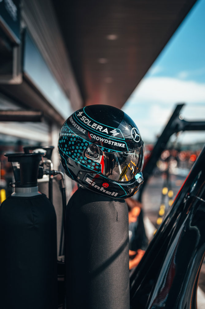 Un casco del team Mercedes-AMG PETRONAS di Formula 1 con i loghi ben visibili degli sponsor, tra cui Einhell, CrowdStrike e Solera, è posizionato su una bombola nella corsia dei box. La visiera riflette l’ambiente del garage.