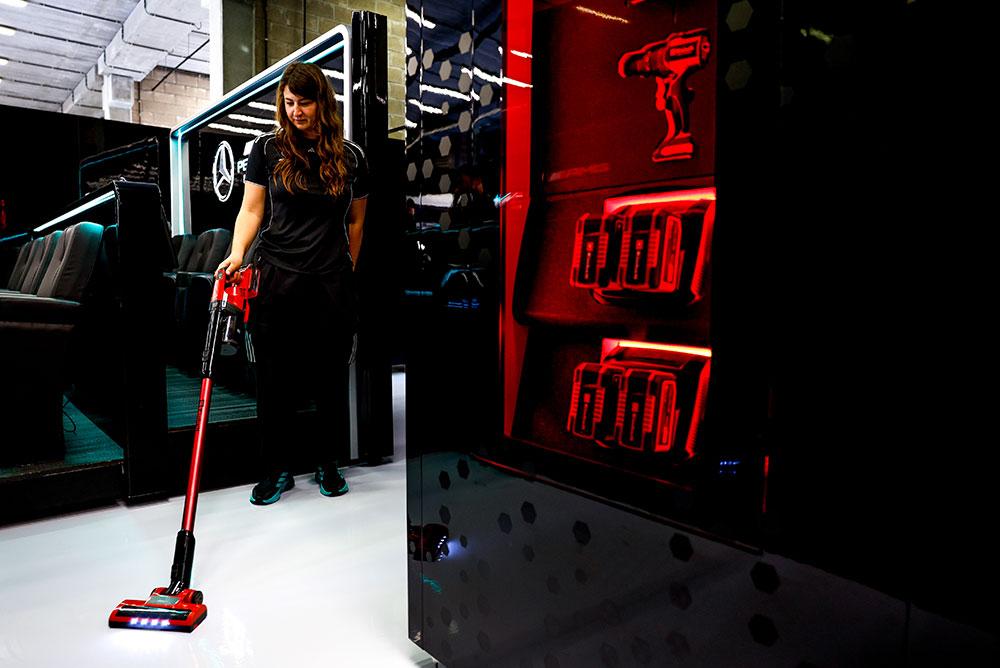 Una donna del team Mercedes-AMG PETRONAS di Formula 1 sta pulendo un pavimento bianco lucido con un aspirapolvere a batteria Einhell. A destra, uno scaffale illuminato di rosso espone diverse batterie Einhell Power X-Change e un trapano a batteria.