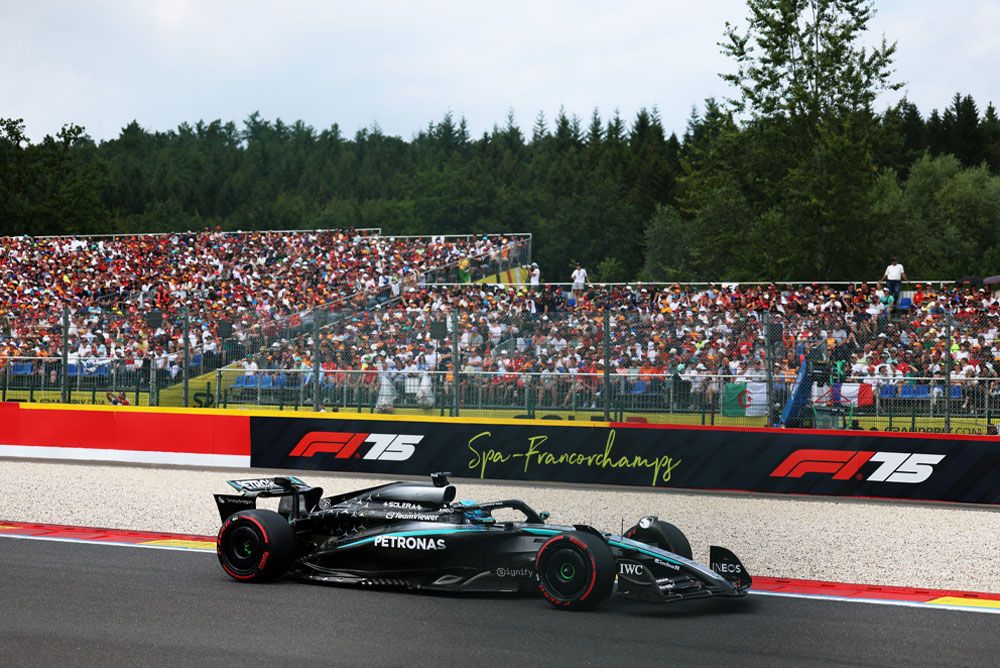 Un’auto di Formula 1 del team Mercedes-AMG PETRONAS sta gareggiando sul circuito di Spa-Francorchamps. Sullo sfondo, le tribune piene di tifosi sventolano bandiere internazionali. Il logo di Spa-Francorchamps è visibile sulla barriera.