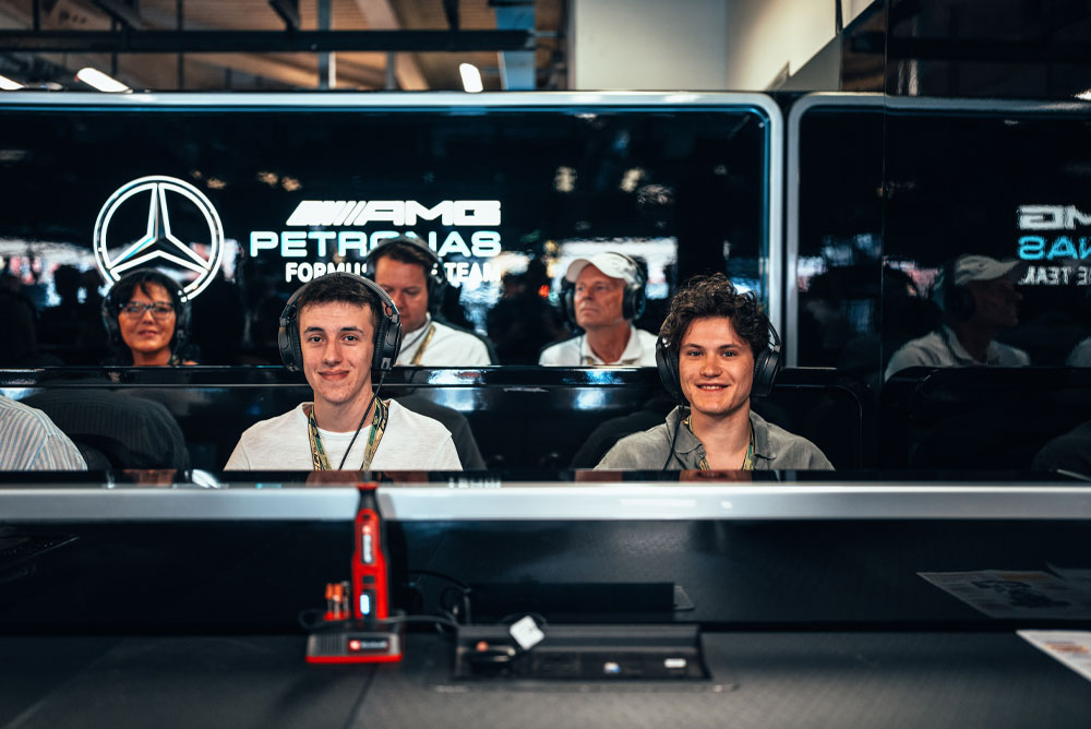 Due uomini sono seduti nel garage del team Mercedes-AMG PETRONAS di Formula 1, indossando cuffie e godendosi l’esperienza dietro le quinte. Un dispositivo rosso Einhell è visibile in primo piano.