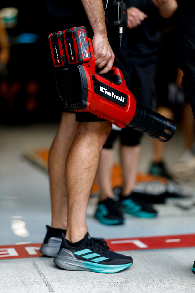 Un membro del team tiene in mano un soffiatore a batteria rosso Einhell con due batterie Power X-Change. La scena si svolge nell'area dei box durante un evento di Formula 1.