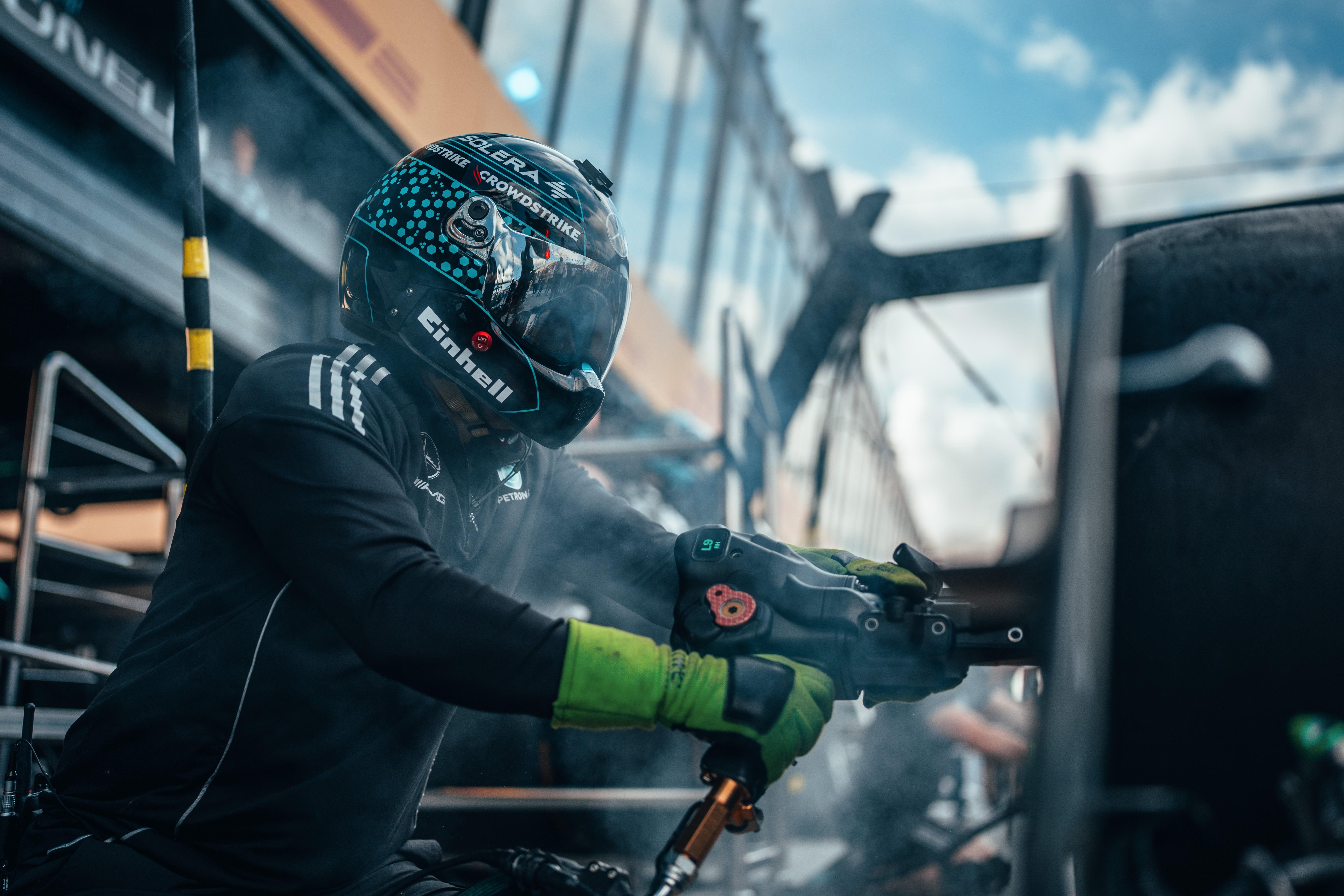 Un membro della squadra ai box di Formula 1, con casco e guanti, utilizza una pistola pneumatica per cambiare le ruote durante un pit stop, circondato da fumo e attrezzi.