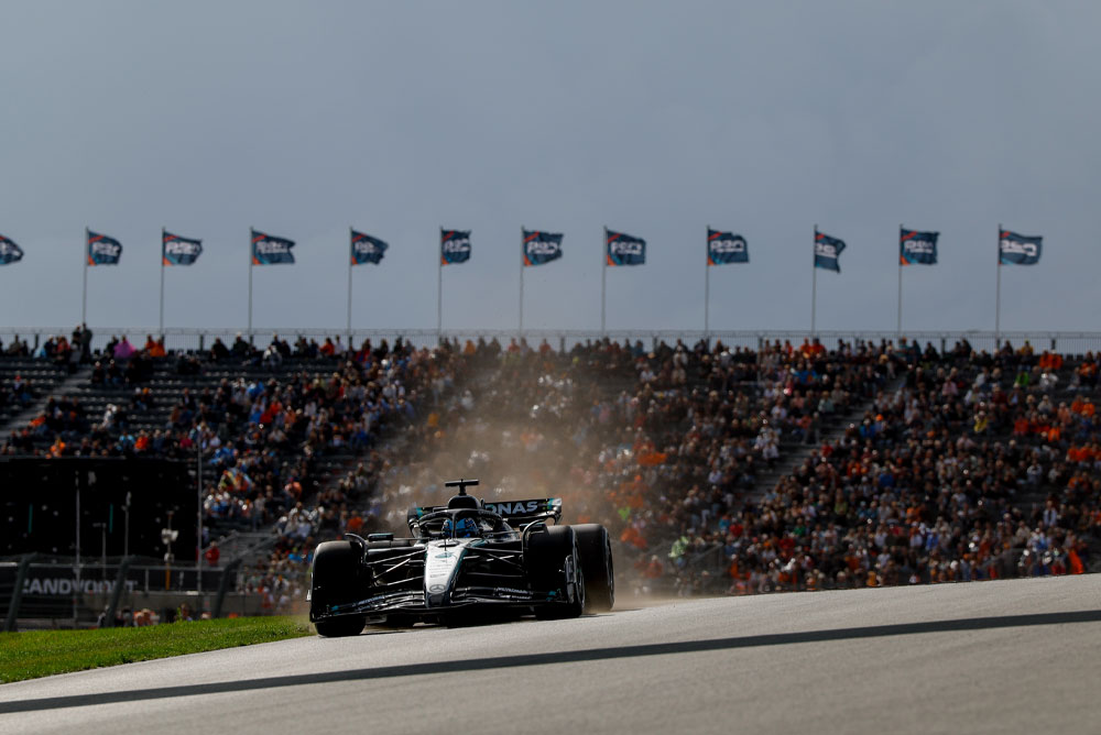 Un’auto di Formula 1 del team Mercedes-AMG PETRONAS sfreccia davanti alle tribune gremite durante una gara. Sullo sfondo, le bandiere ondeggiano creando un’atmosfera vibrante.