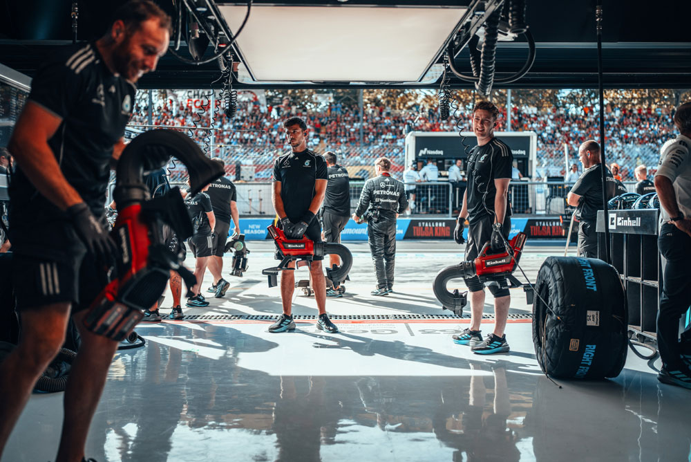 I meccanici del team Mercedes-AMG PETRONAS di Formula 1 si preparano nel box con soffiatori a batteria Einhell prima di un pit stop. Sullo sfondo, le tribune sono piene di tifosi.
