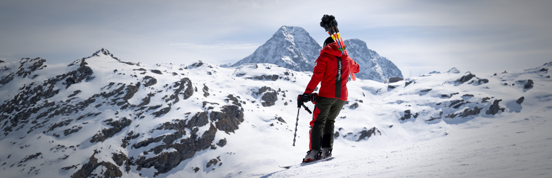 Ein Swiss-Ski-Team-Mitglied mit Slalomstangen auf dem Rücken und einem Einhell Akkubohrschrauber steht auf einem verschneiten Hang mit weitem Blick auf die Alpen.