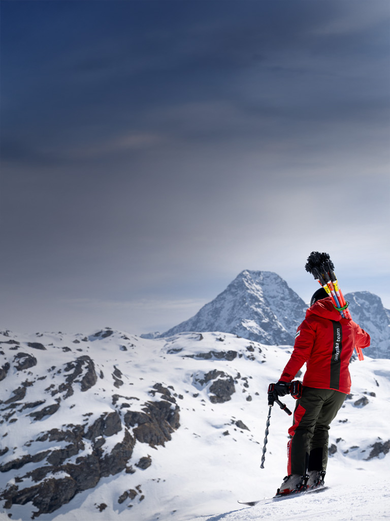 Ein Swiss-Ski-Team-Mitglied mit Slalomstangen auf dem Rücken und einem Einhell Akkubohrschrauber steht auf einem verschneiten Hang mit weitem Blick auf die Alpen.