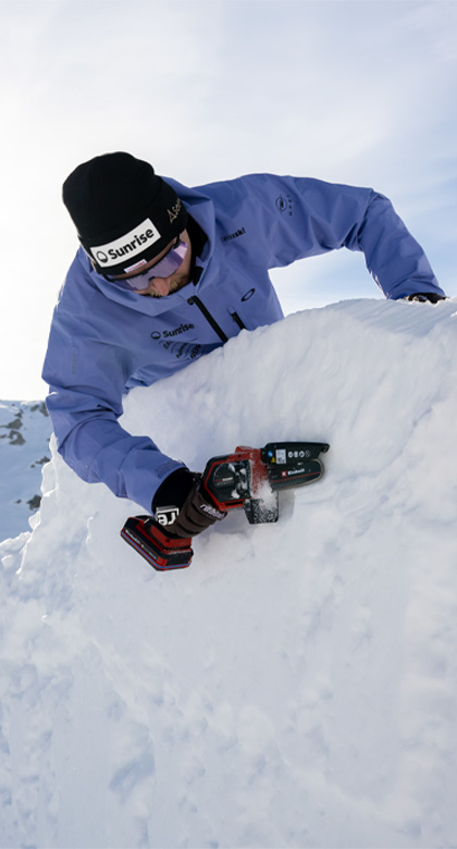 Ein Swiss-Ski-Team-Mitglied in hellblauer Jacke arbeitet mit einer kleinen Einhell Akku-Kettensäge an einer Schneewand.