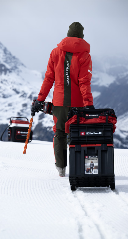 Ein Swiss-Ski-Team-Mitglied zieht einen großen schwarzen Einhell-Werkzeugkoffer auf Schneefläche und hält einen Einhell Akkubohrschrauber mit orangem Bohrer.