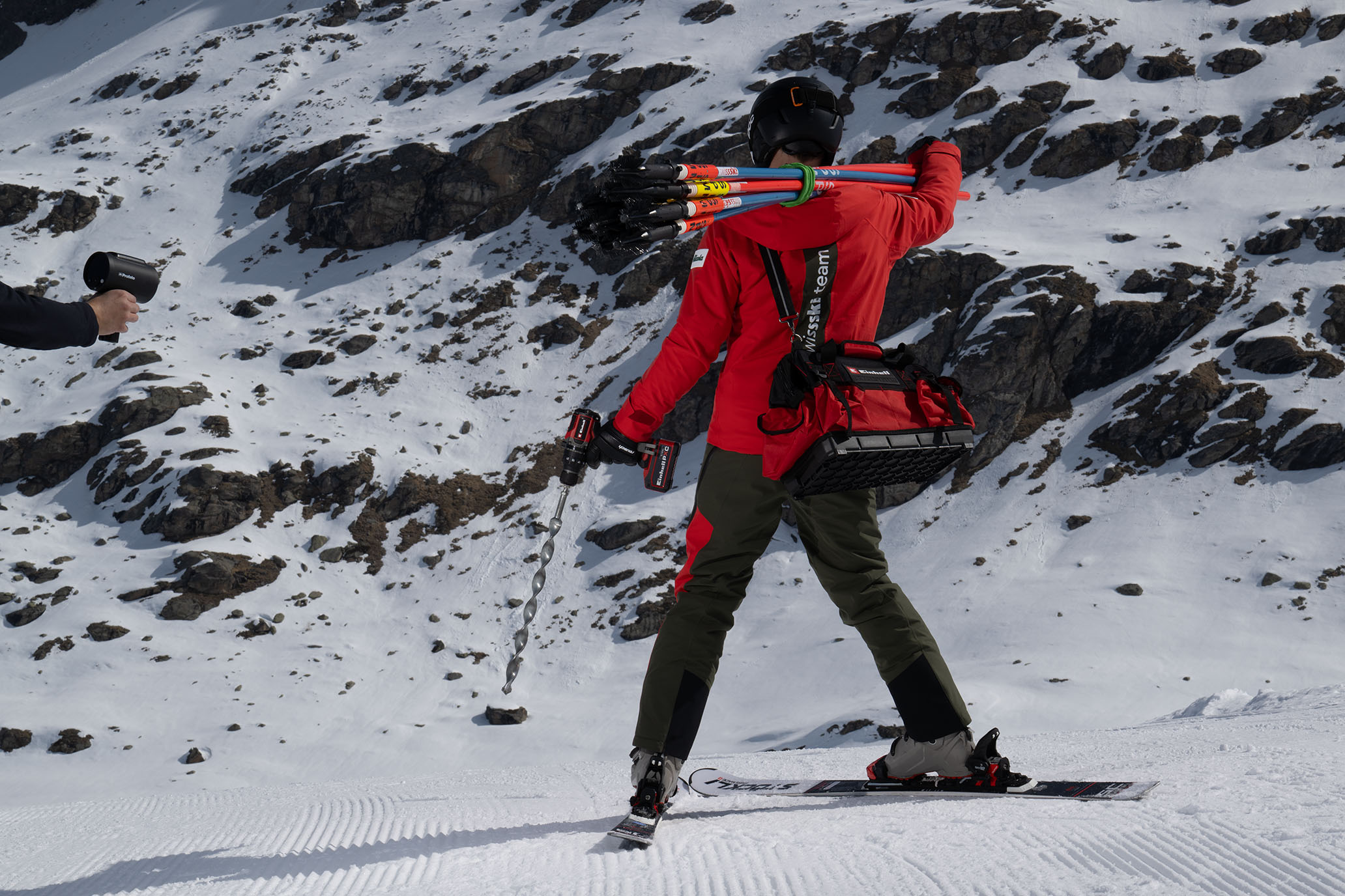 Ein Swiss-Ski-Team-Mitglied mit Slalomstangen und einem roten Werkzeugträger bohrt mit einem Einhell Akkubohrschrauber in den Schnee.