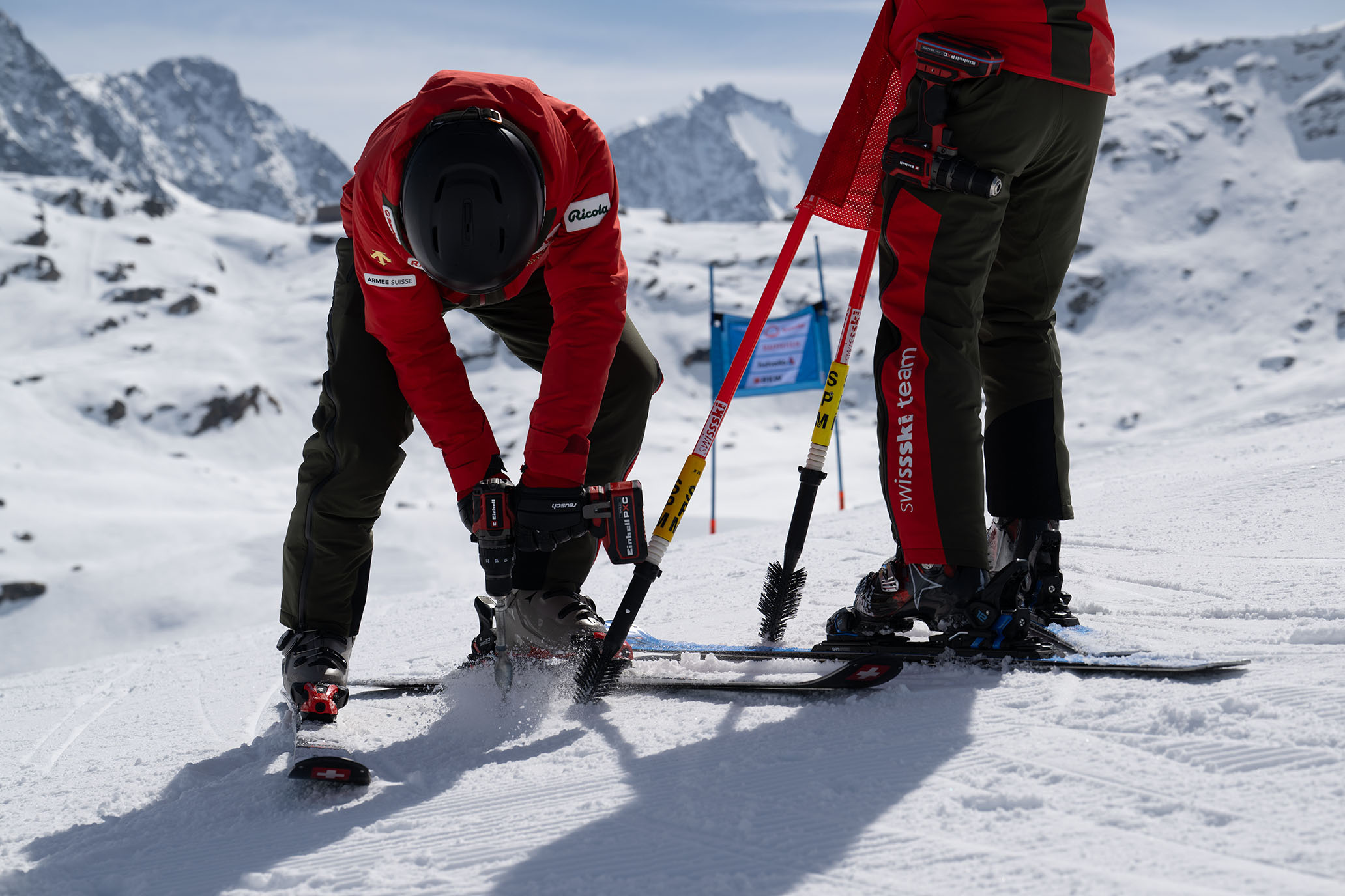 Zwei Swiss-Ski-Team-Mitglieder arbeiten im Schnee: einer bohrt mit dem Einhell Akkubohrschrauber, der andere hält Slalomstangen.
