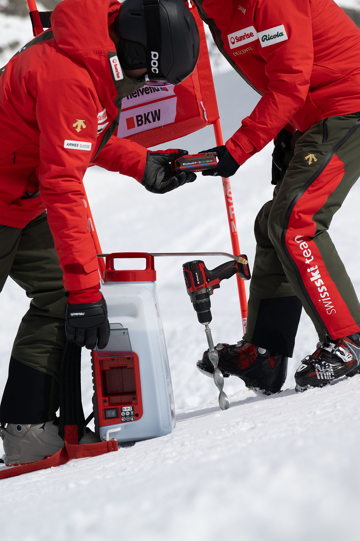 Zwei Swiss-Ski-Team-Mitglieder setzen einen Power X-Change Akku in ein weißes Einhell-Gerät ein, daneben ein Akkubohrschrauber mit Eisbohrer.