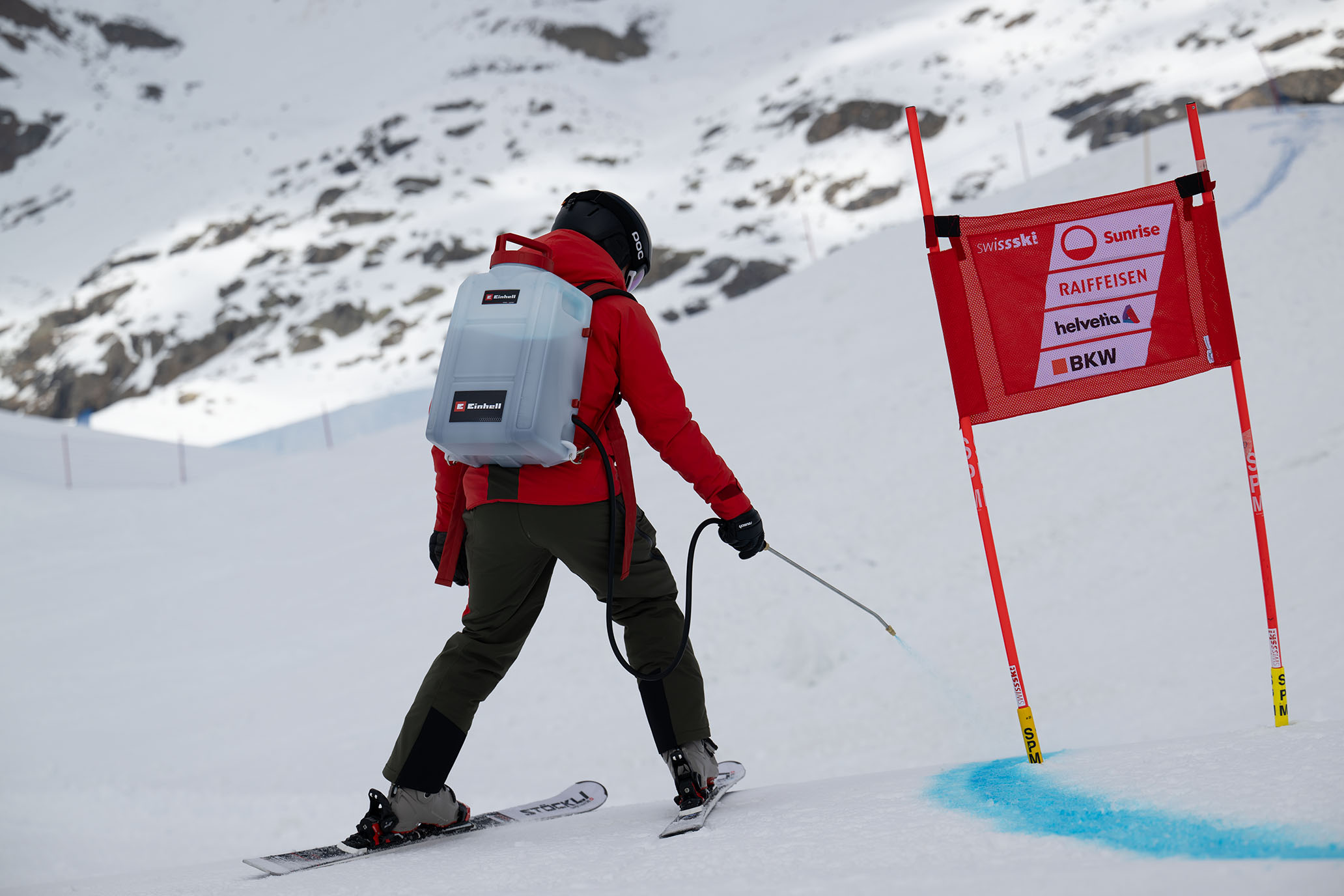 Ein Swiss-Ski-Team-Mitglied markiert mit einem weißen Einhell-Rucksack-Sprühsystem eine blaue Linie im Schnee an einem Slalomtor.