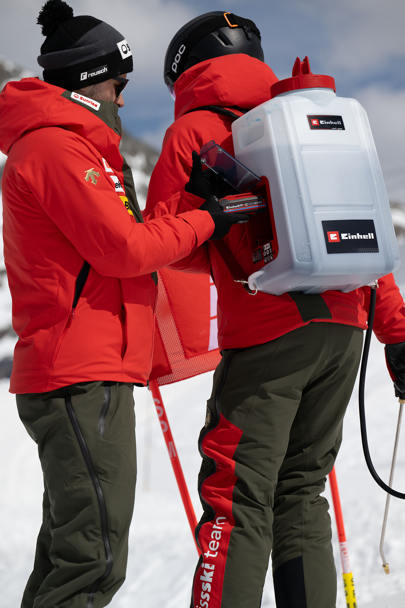 Ein Swiss-Ski-Team-Mitglied trägt einen weißen Einhell-Rucksack mit Flüssigkeitstank, während ein Kollege einen Power X-Change Akku einsetzt.