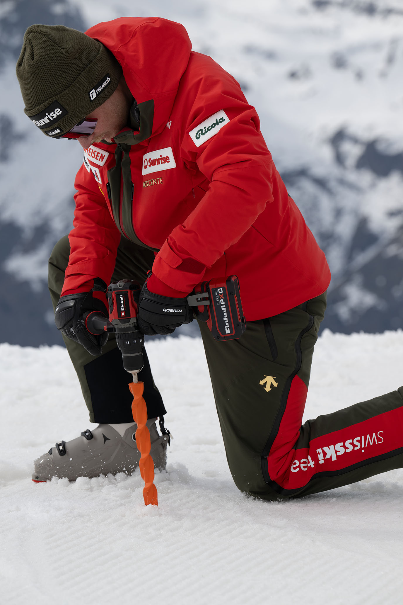 Ein Swiss-Ski-Team-Mitglied kniet auf dem Schnee und bohrt mit einem Einhell Akkubohrschrauber und orangefarbenem Bohrer ein Loch.