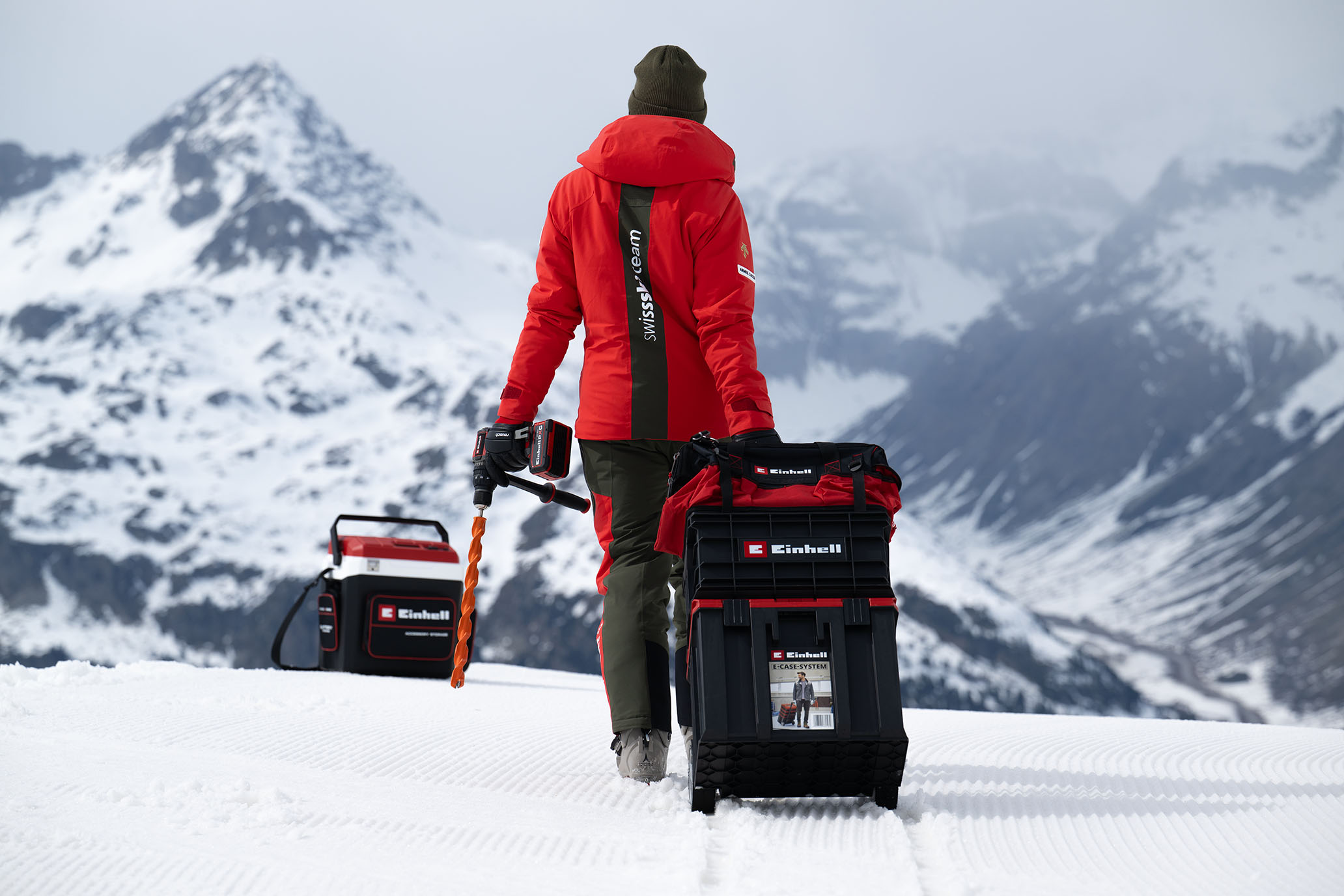 Ein Swiss-Ski-Team-Mitglied zieht einen großen schwarzen Einhell-Werkzeugkoffer auf Schneefläche und hält einen Einhell Akkubohrschrauber mit orangem Bohrer.