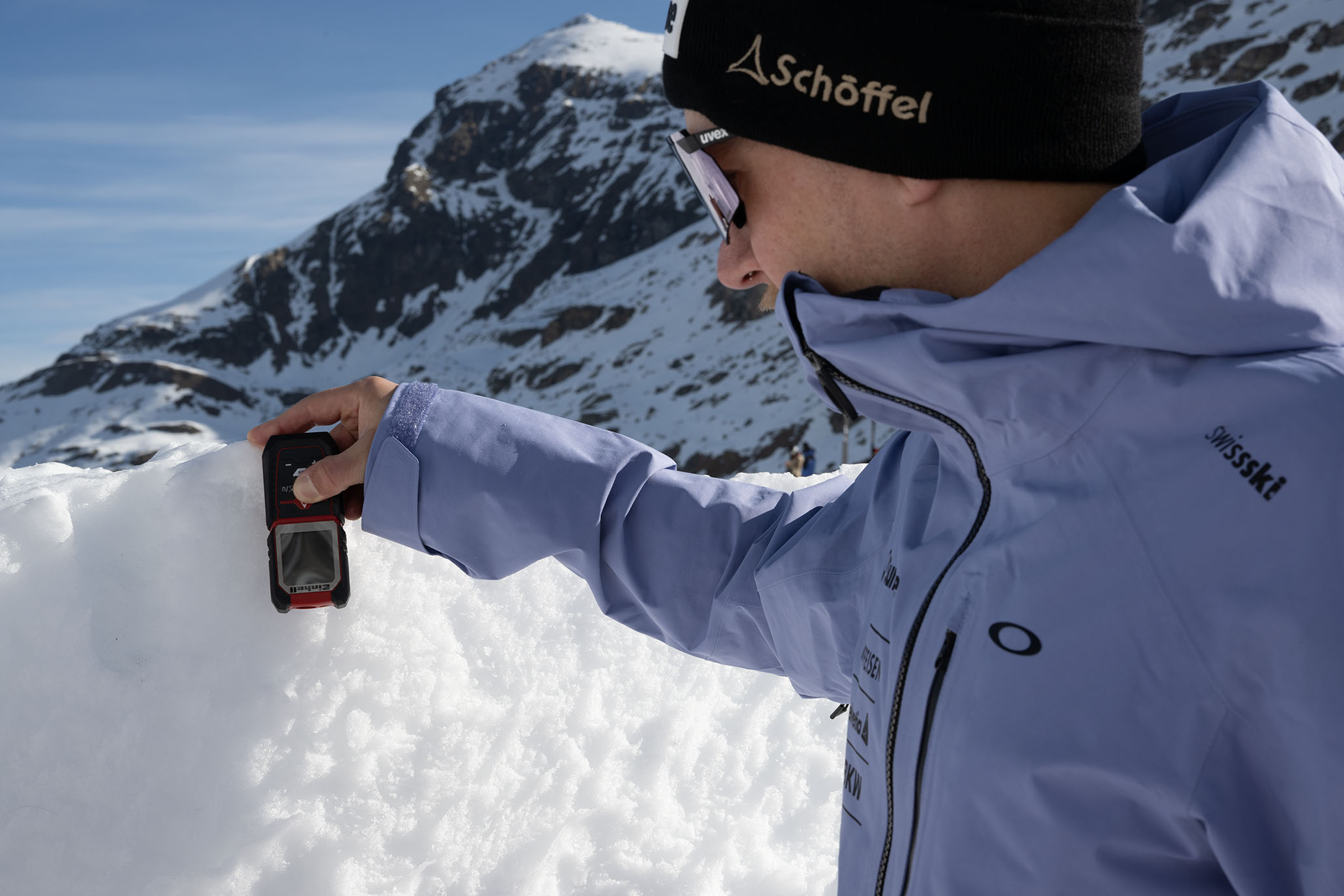 Ein Swiss-Ski-Team-Mitglied in hellblauer Jacke misst mit einem roten Einhell-Laser-Entfernungsmesser eine Schneehöhe.