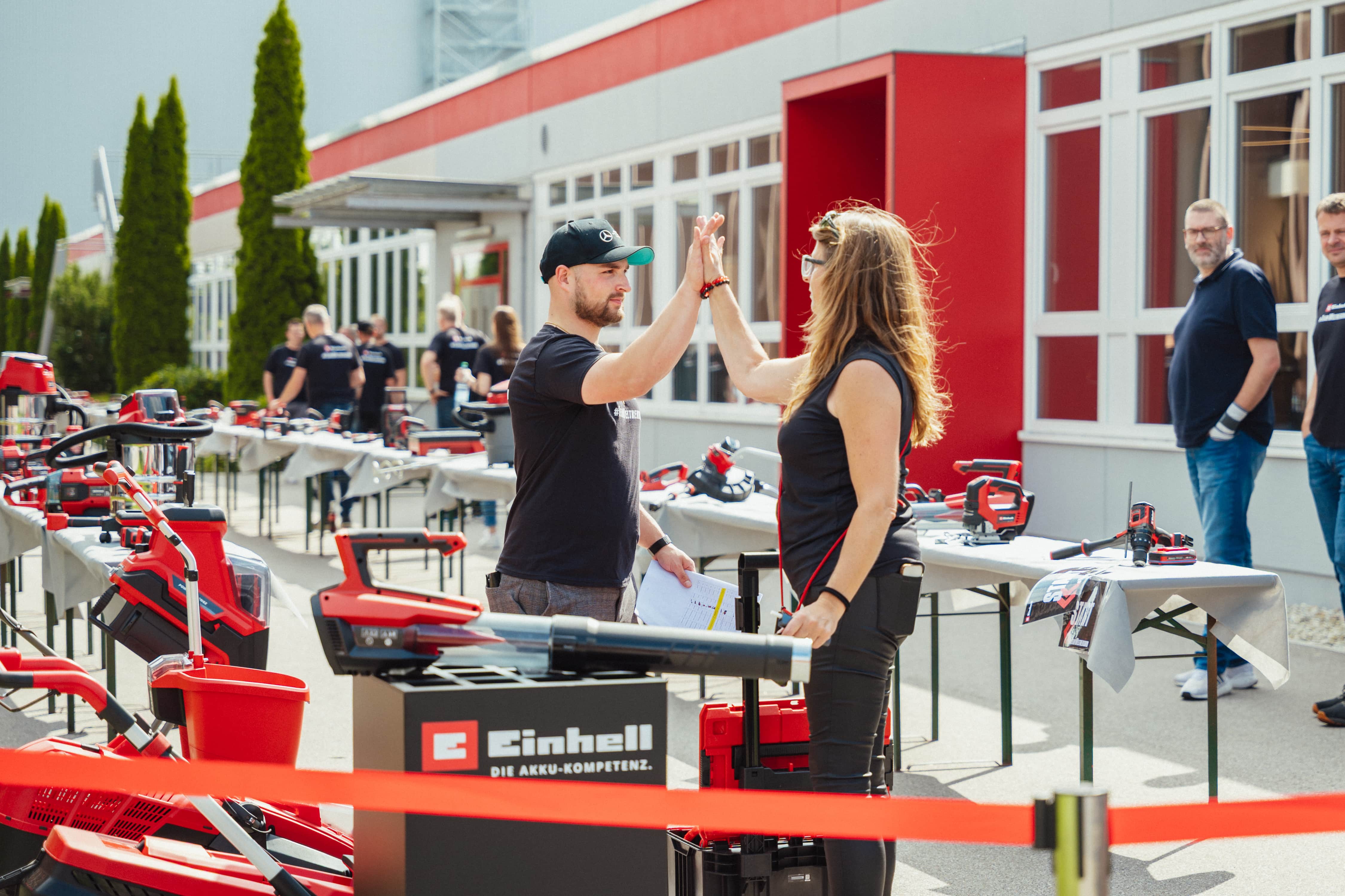 Ein Mann und eine Frau geben sich ein High-Five zwischen aufgebauten Einhell-Geräten im Außenbereich.