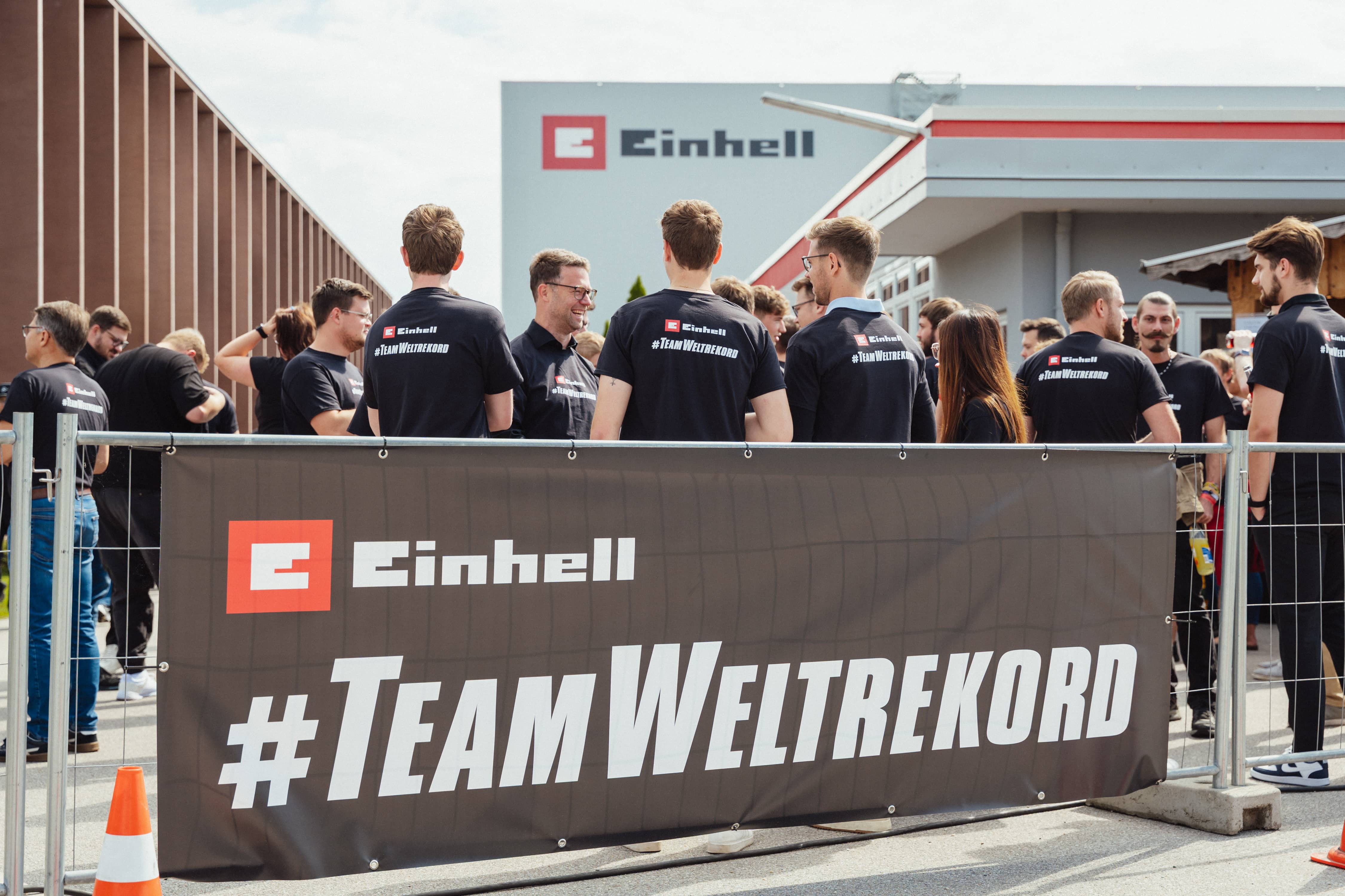 Teilnehmer stehen hinter einem Zaun mit großem Banner „Einhell #TeamWeltrekord“ auf dem Werksgelände.
