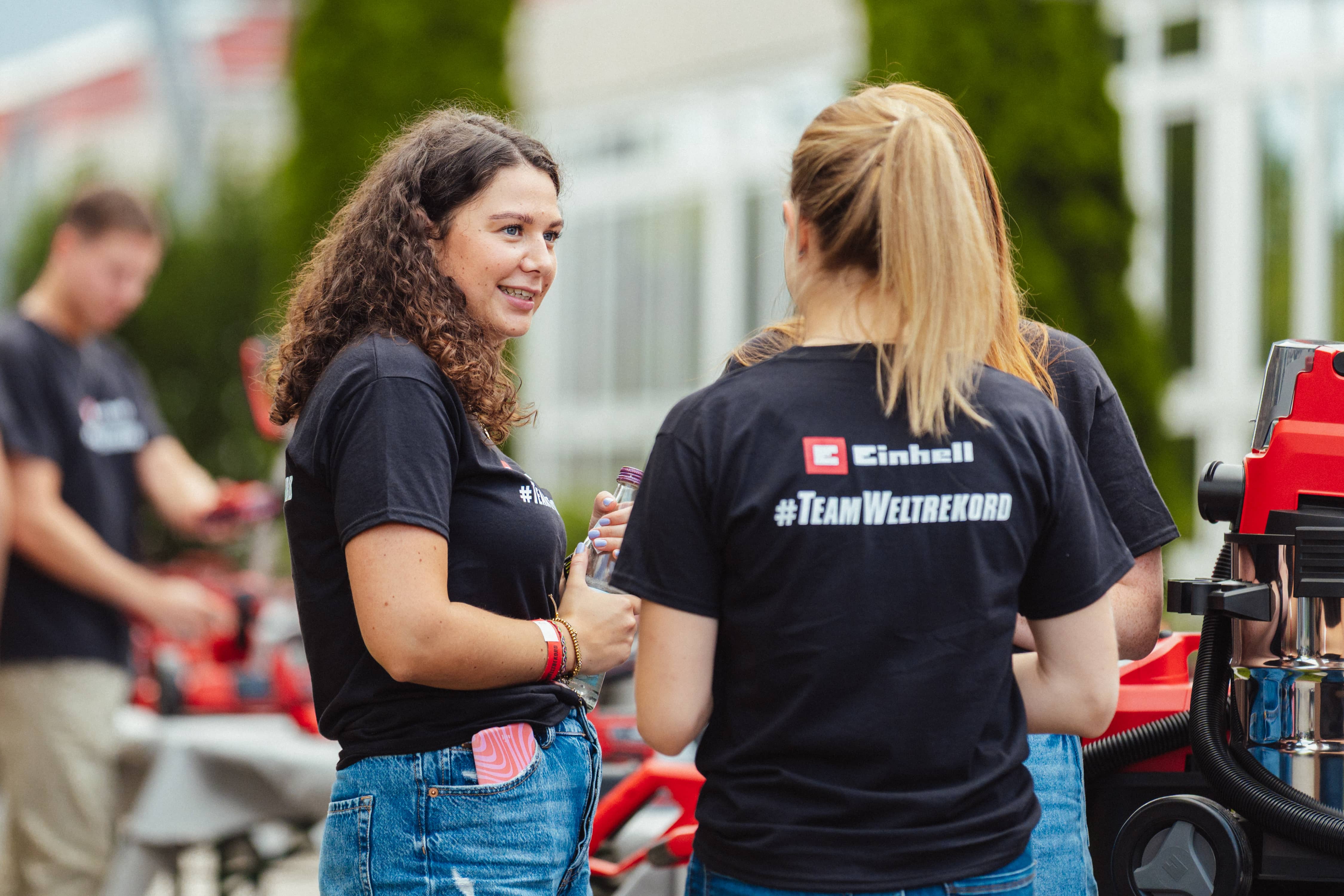 Zwei junge Frauen in "#TeamWeltrekord"-Shirts unterhalten sich vor Einhell-Geräten im Freien.