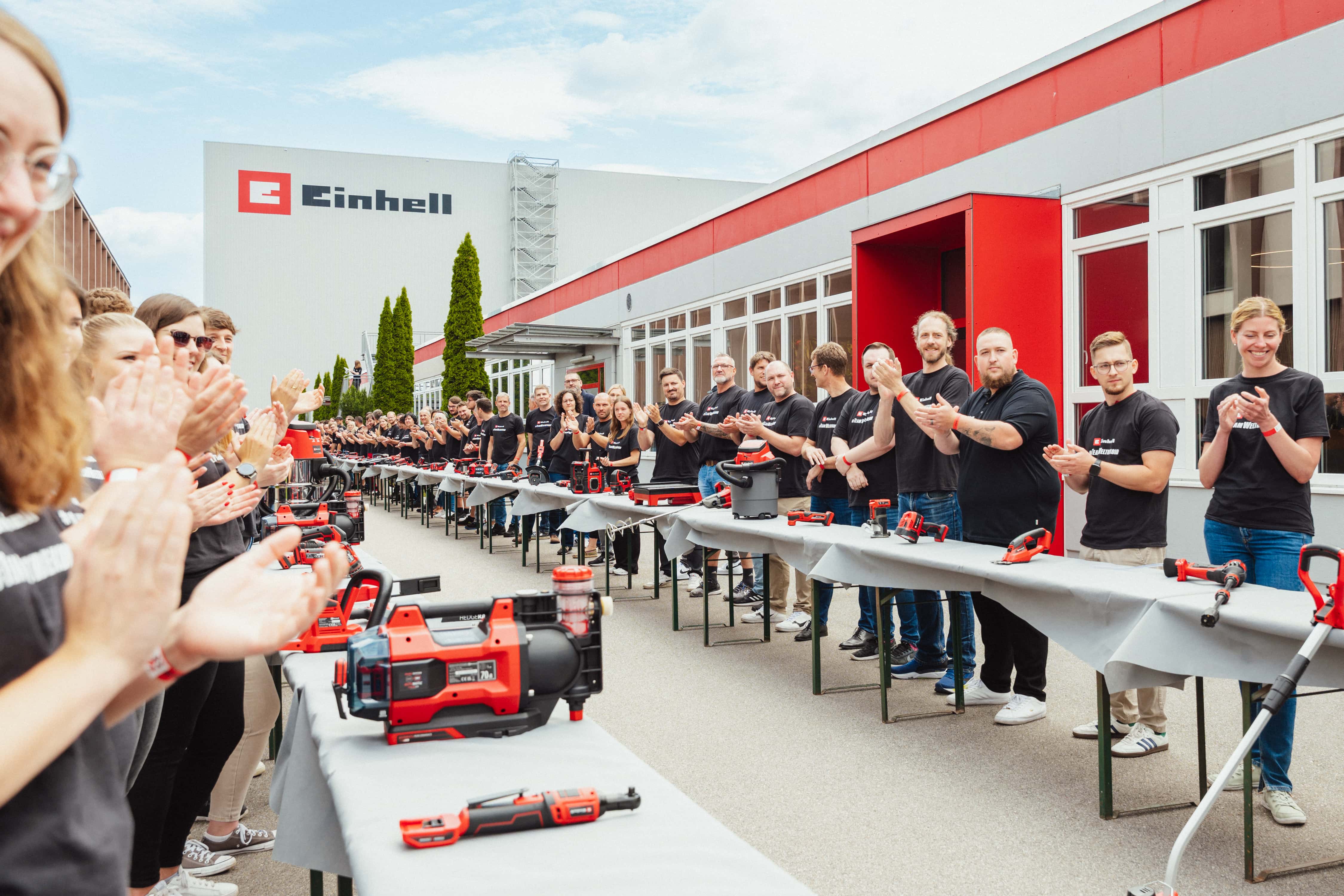 Zahlreiche Einhell-Mitarbeitende klatschen entlang einer langen Tischreihe mit Akku-Geräten auf dem Werksgelände.