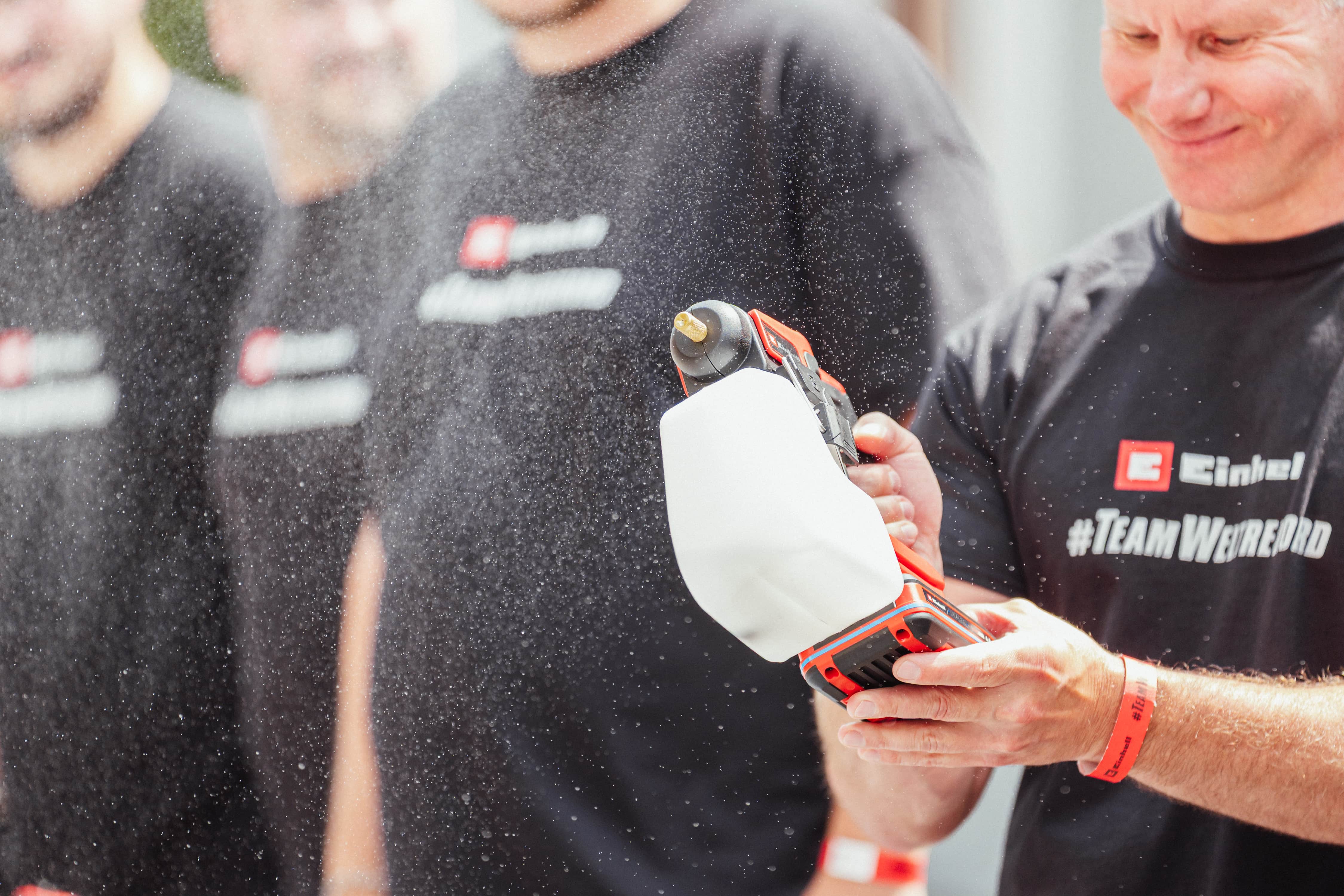 Ein Mann mit "#TeamWeltrekord"-Shirt hält eine Einhell Akku-Sprühsystem, aus dem feiner Sprühnebel austritt.