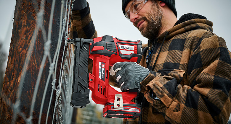 A man is using an Einhell cordless stapler FIXETTO to attach wire to a fence, wearing gloves and safety glasses.