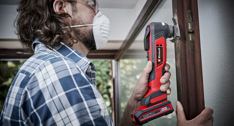 A person wearing safety glasses and a face mask uses a Einhell multi-tool to work on a window frame.