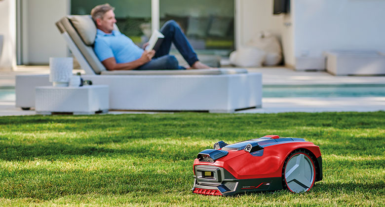 Ein Mann entspannt auf einer Liege im Garten, vor ihm mäht ein Mähroboter den Rasen.