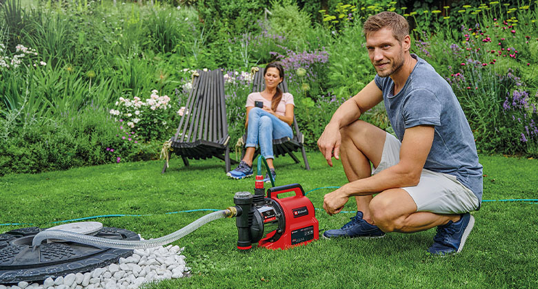 Ein Mann kniend im Garten neben einer Gartenpumpe, die an einen unterirdischen Wassertank angeschlossen ist. Im Hintergrund sitzt eine Frau im Gartenstuhl.